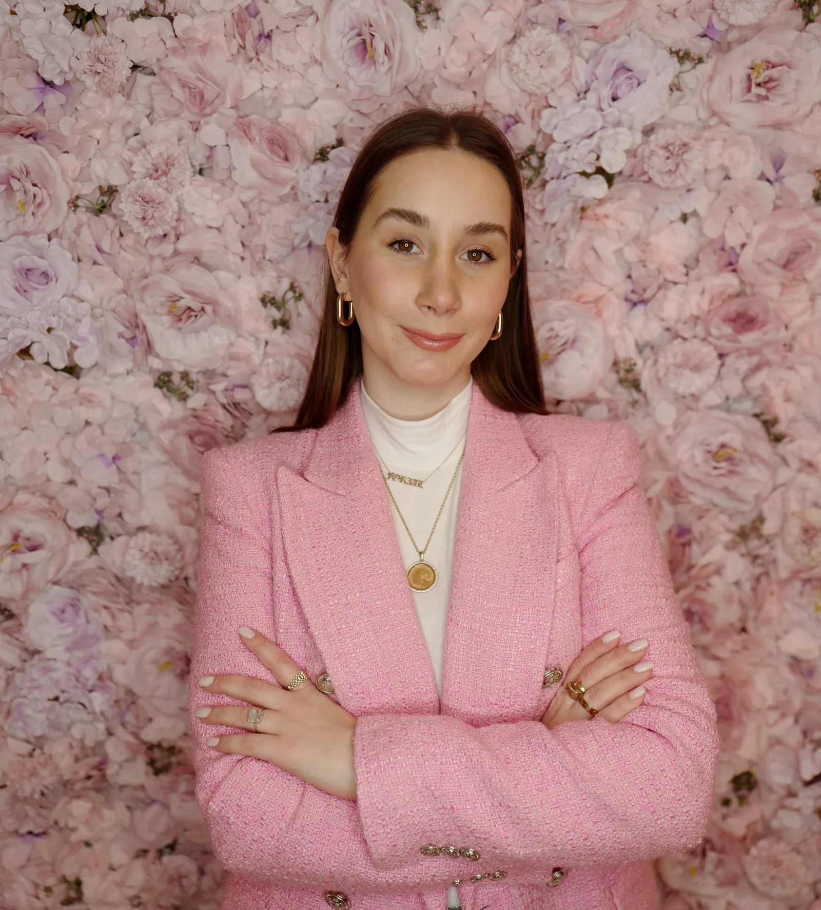 Aleksandra i rosa dressjakke, med armene i kors foran en vegg av lyserosa roser