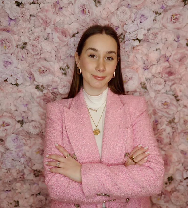 Aleksandra i rosa dressjakke, med armene i kors foran en vegg av lyserosa roser