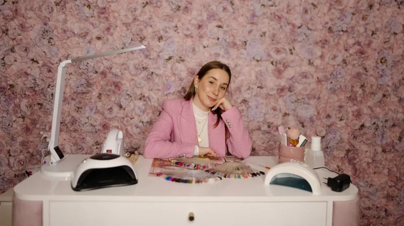 Aleksandra sitter ved arbeidsbordet sitt med neglelakk og lampe, og smiler mot kamera